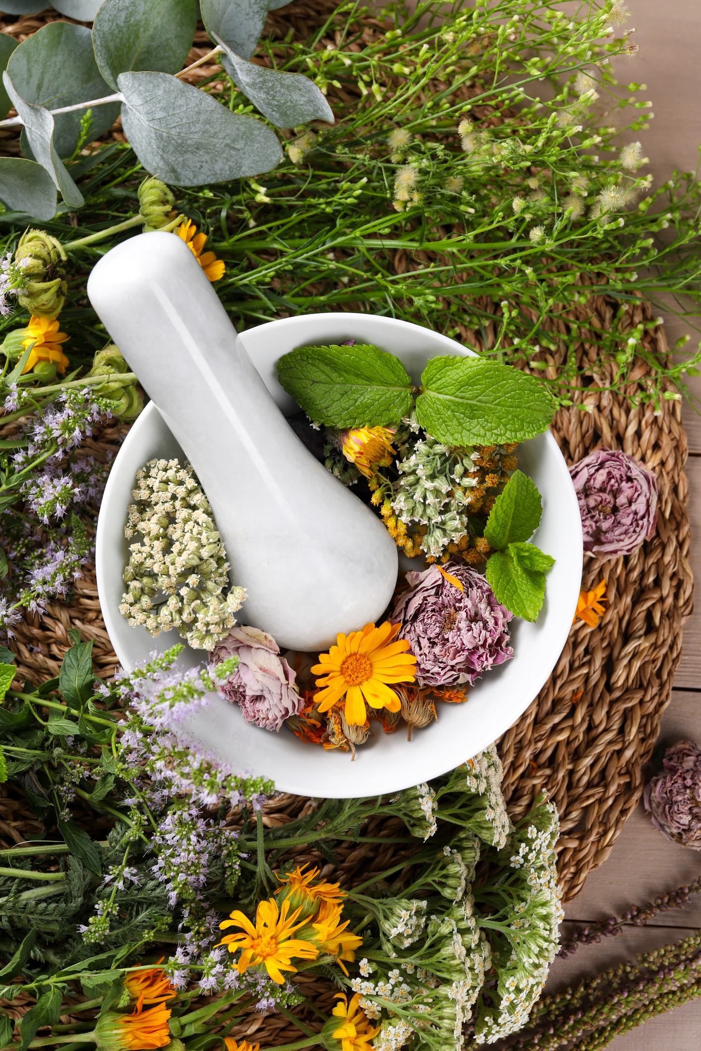 A white mortar and pestle filled with assorted dried flowers and fresh mint leaves, surrounded by various fresh herbs and greenery on a woven mat.