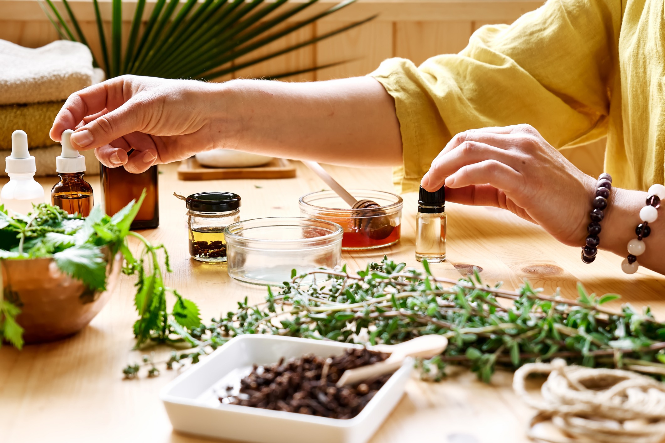 A person in a yellow shirt prepares natural remedies at a wooden table, surrounded by glass jars, essential oil bottles, bowls of herbs, honey, and fresh green plants.