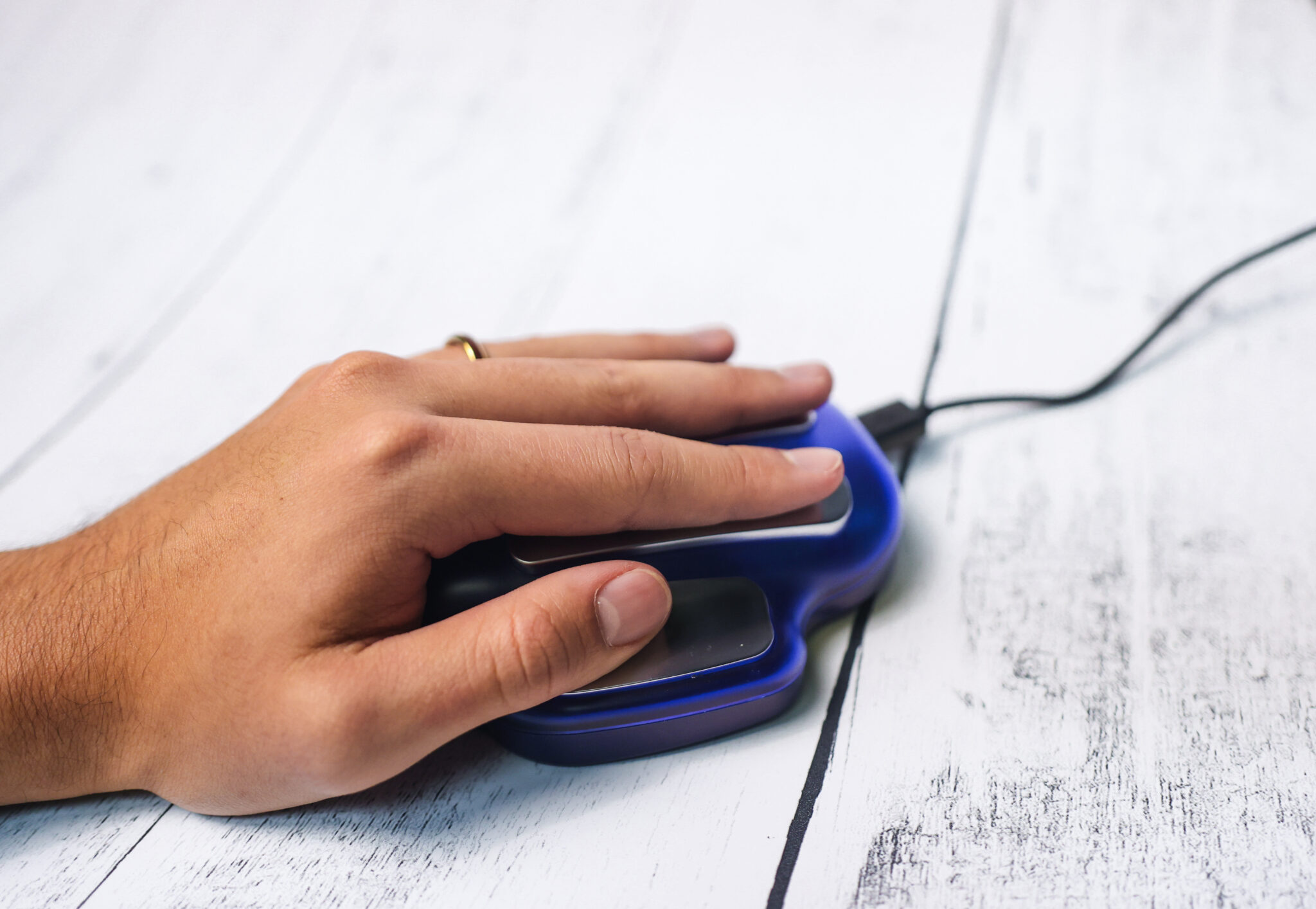 A hand with a gold ring rests on an ergonomic vertical computer mouse placed on a white wooden surface.